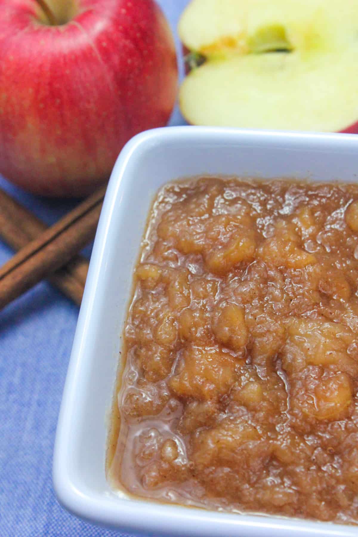 A bowl of Slow Cooker Applesauce with apples and cinnamon sticks next to it on a table.