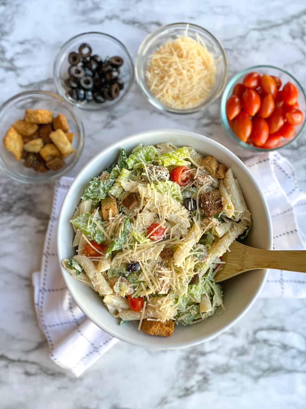 Caesar Pasta Salad in bowl with spoon and croutons, olives, cheese, and tomatoes in small bowls in the background