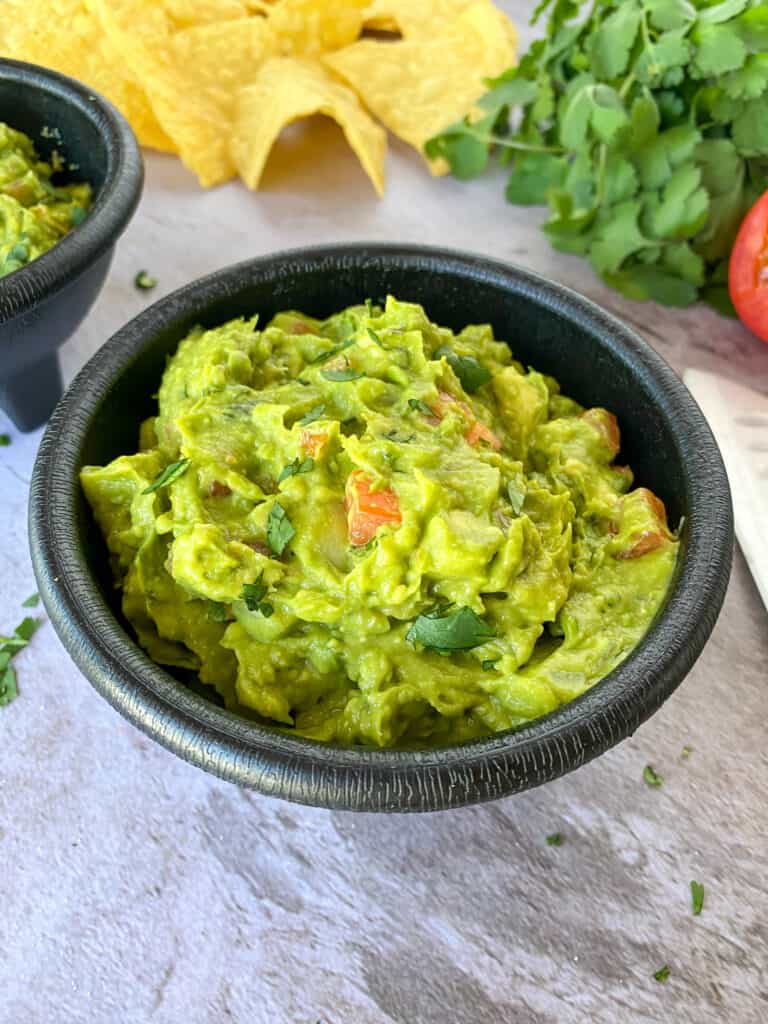 bowl of The Best Easy Homemade Guacamole with chips, cilantro, a tomato, and a chef's knife in the background