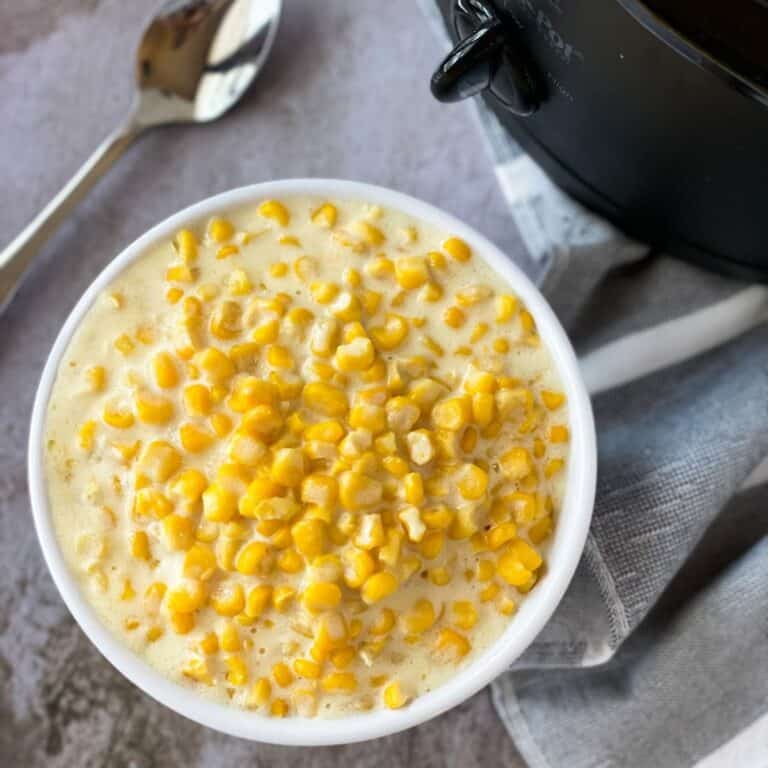 bowl of 5 ingredient creamed corn with a slow cooker, towel, and spoon on the the table