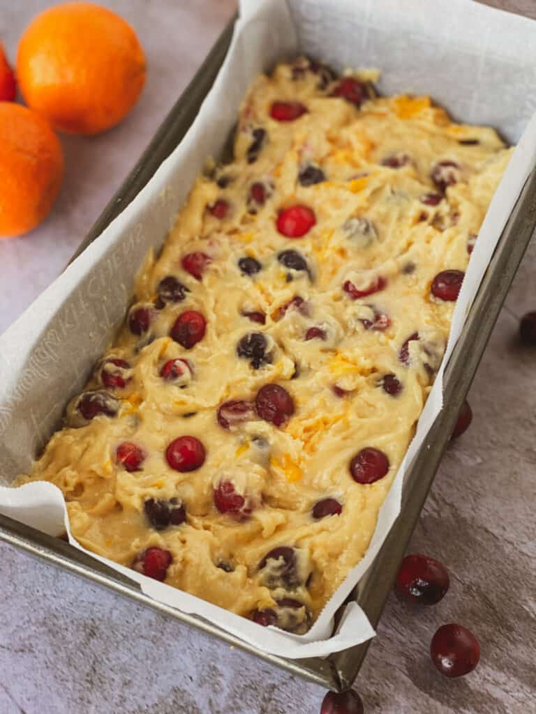 batter in a loaf pan with oranges and cranberries on the table
