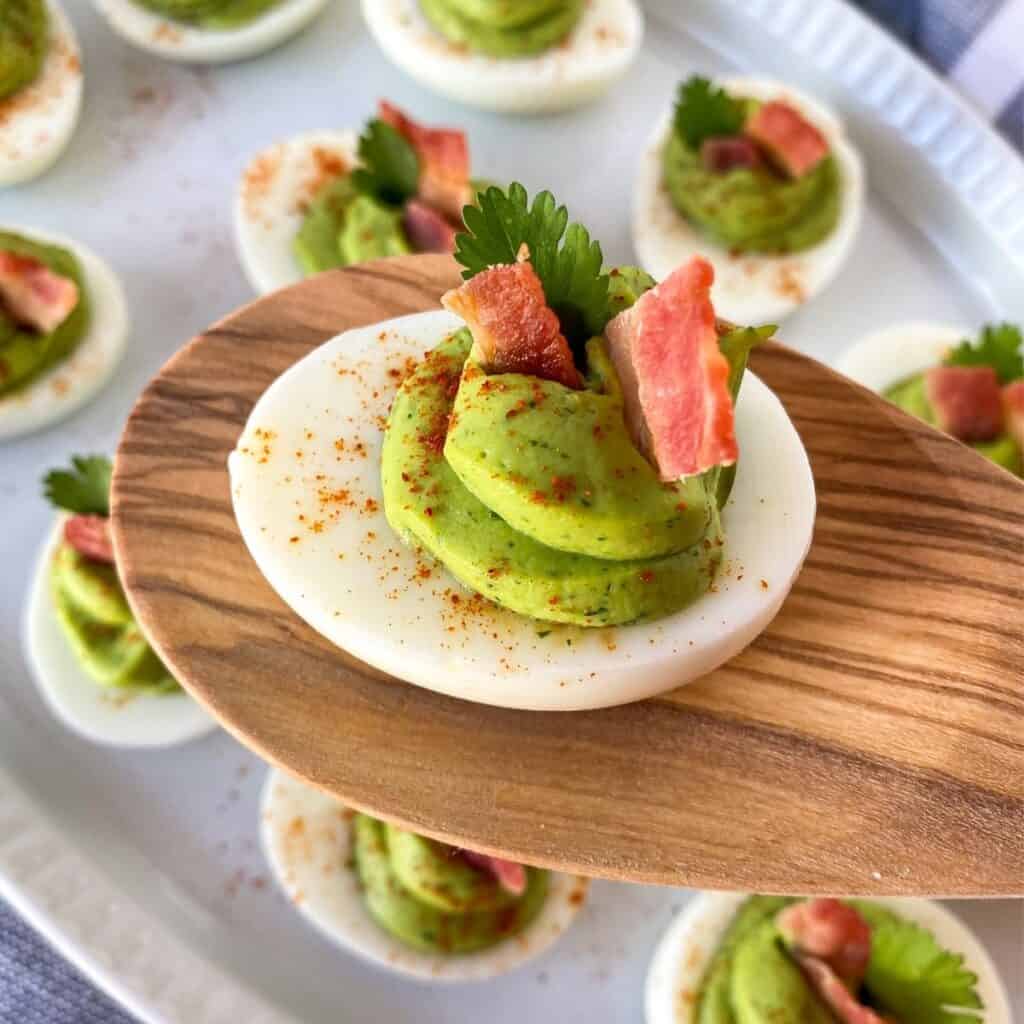 easy bacon avocado deviled egg on a wooden spoon with a plate of eggs in the background