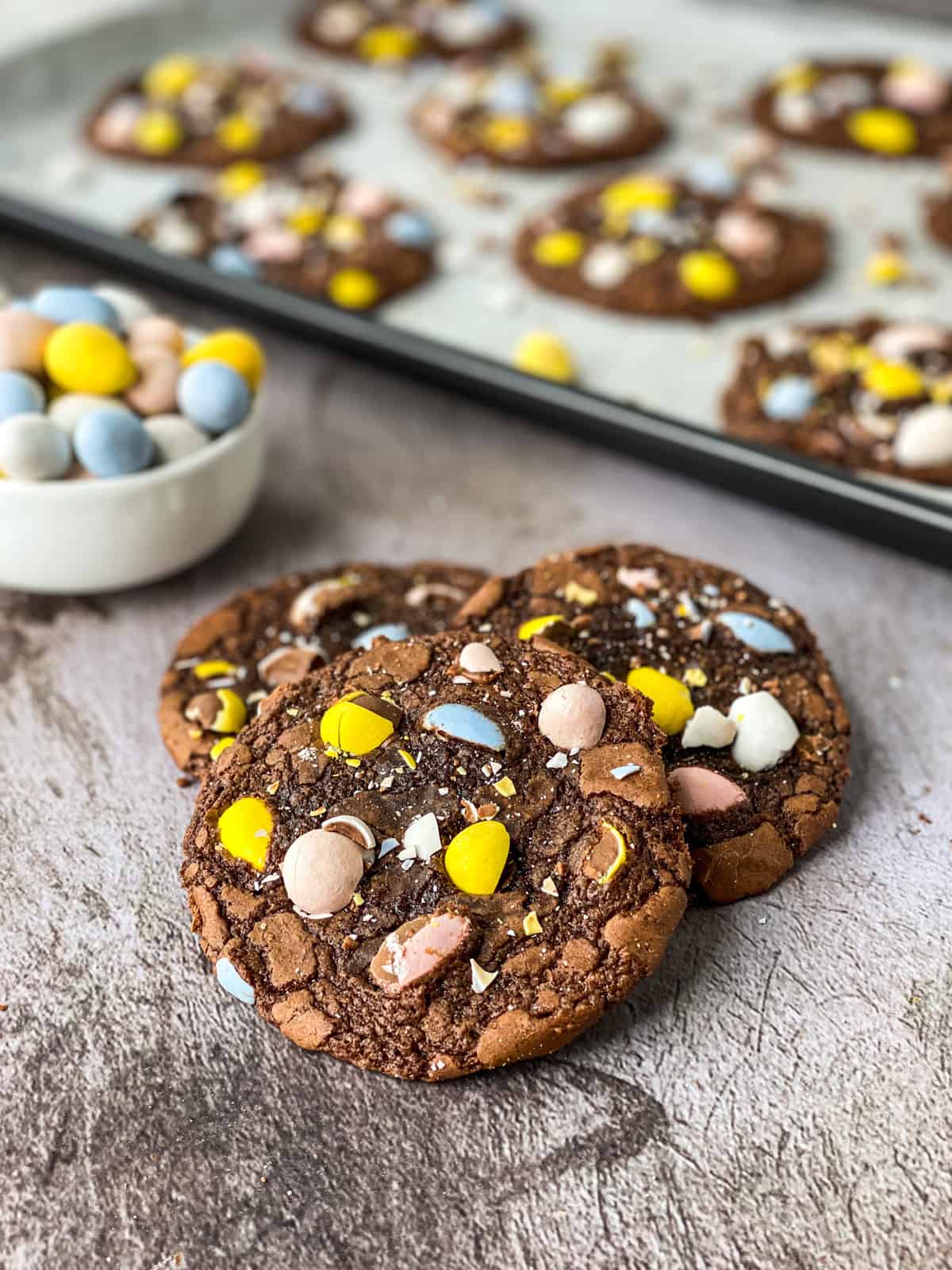 3 cookies on a table with a bowl of cadbury mini eggs and a baking sheet full of cookies