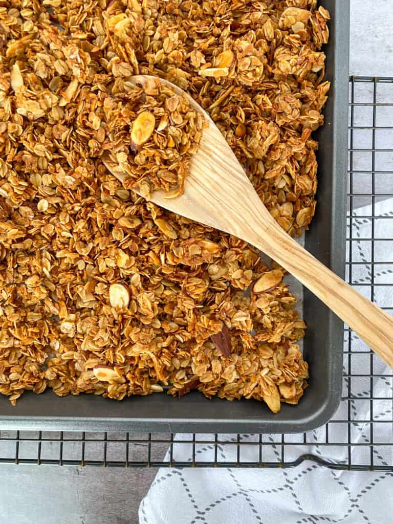 pan of granola with a wooden spoon in it and a cooling rack and cloth napkin in the background