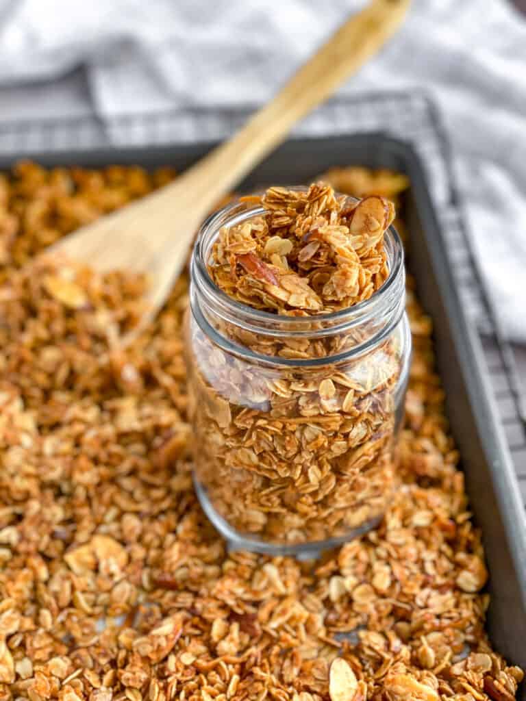 close up of granola in a clear jar on a pan with granola and a wooden spoon and cooling rack in the background