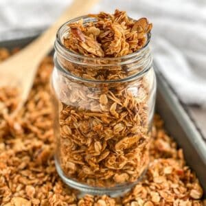 close up of granola in a clear jar on a pan with granola and a wooden spoon in the background