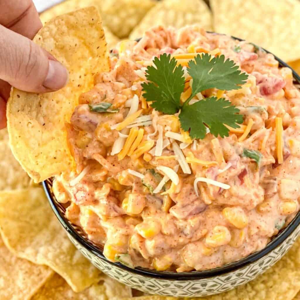a hand scooping the corn dip in a bowl with a tortilla chip