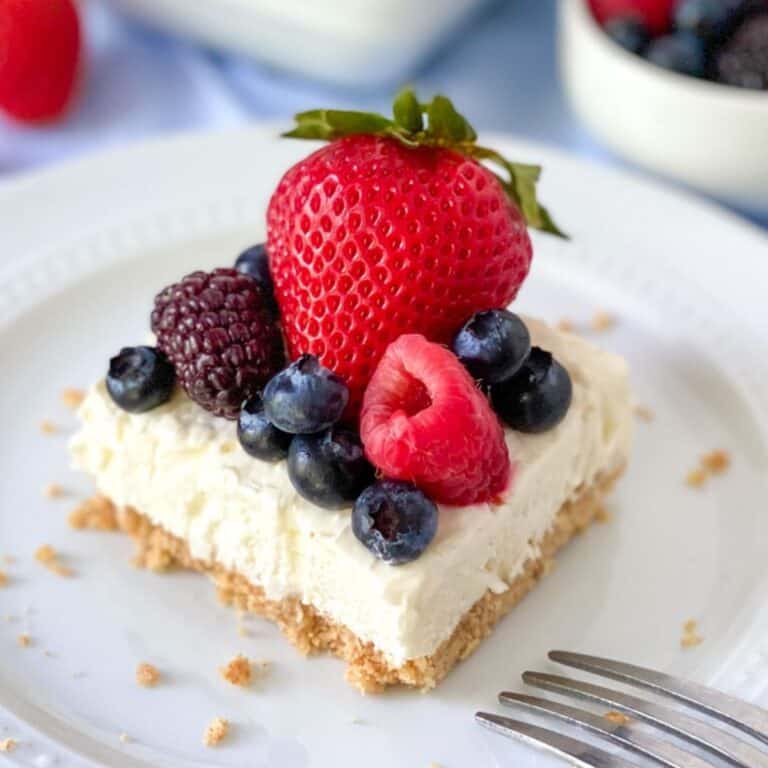 cheesecake bar covered in berries on a plate with a fork, berries in the background, and the pan of cheesecake in the background