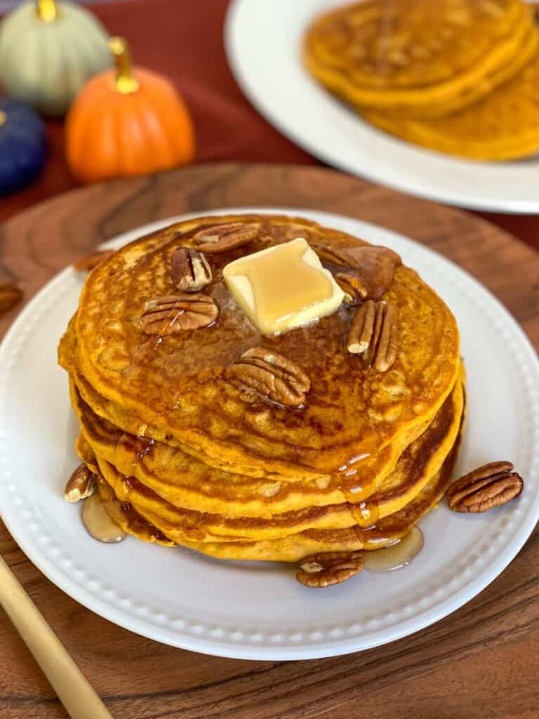 pumpkin spice pancakes with pancake mix on a plate with butter, syrup, and pecans on top and more pancakes on a plate in the background