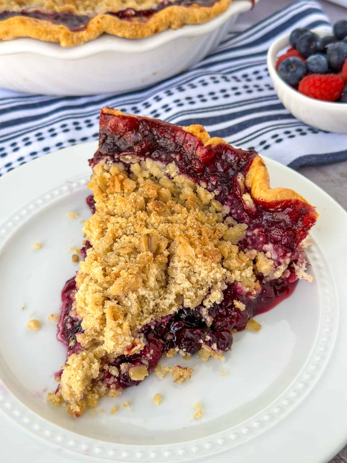 slice of mixed berry pie with oatmeal crumb topping on a plate with a pie in the background and a bowl of berries