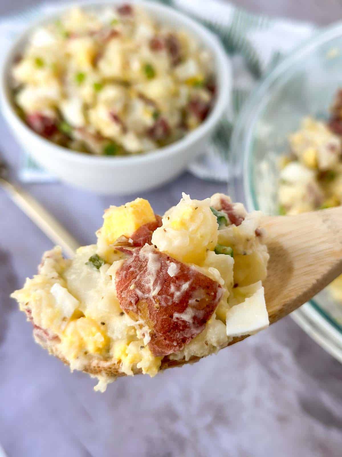 potato salad on a wooden spoon with more in a large bowl and small bowl in the background