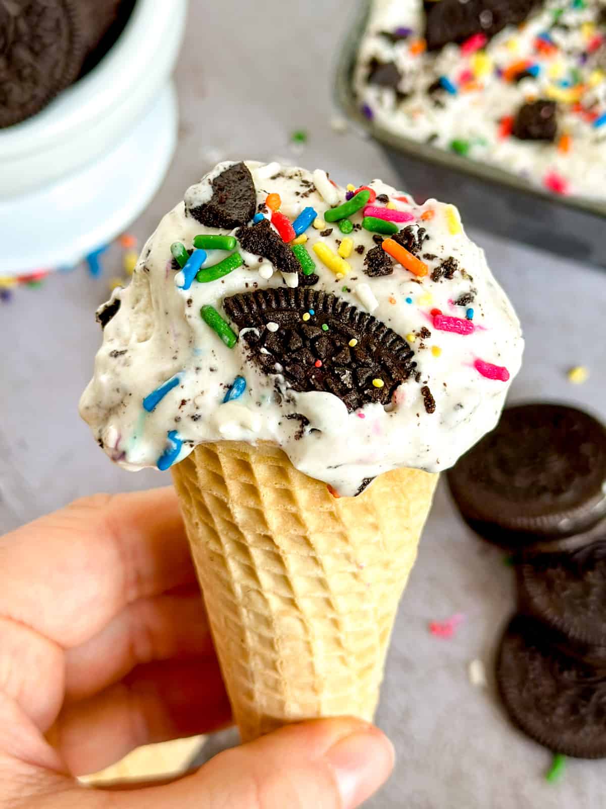 close up of a ice cream cone in a hand with oreos and more ice cream in the backgroun