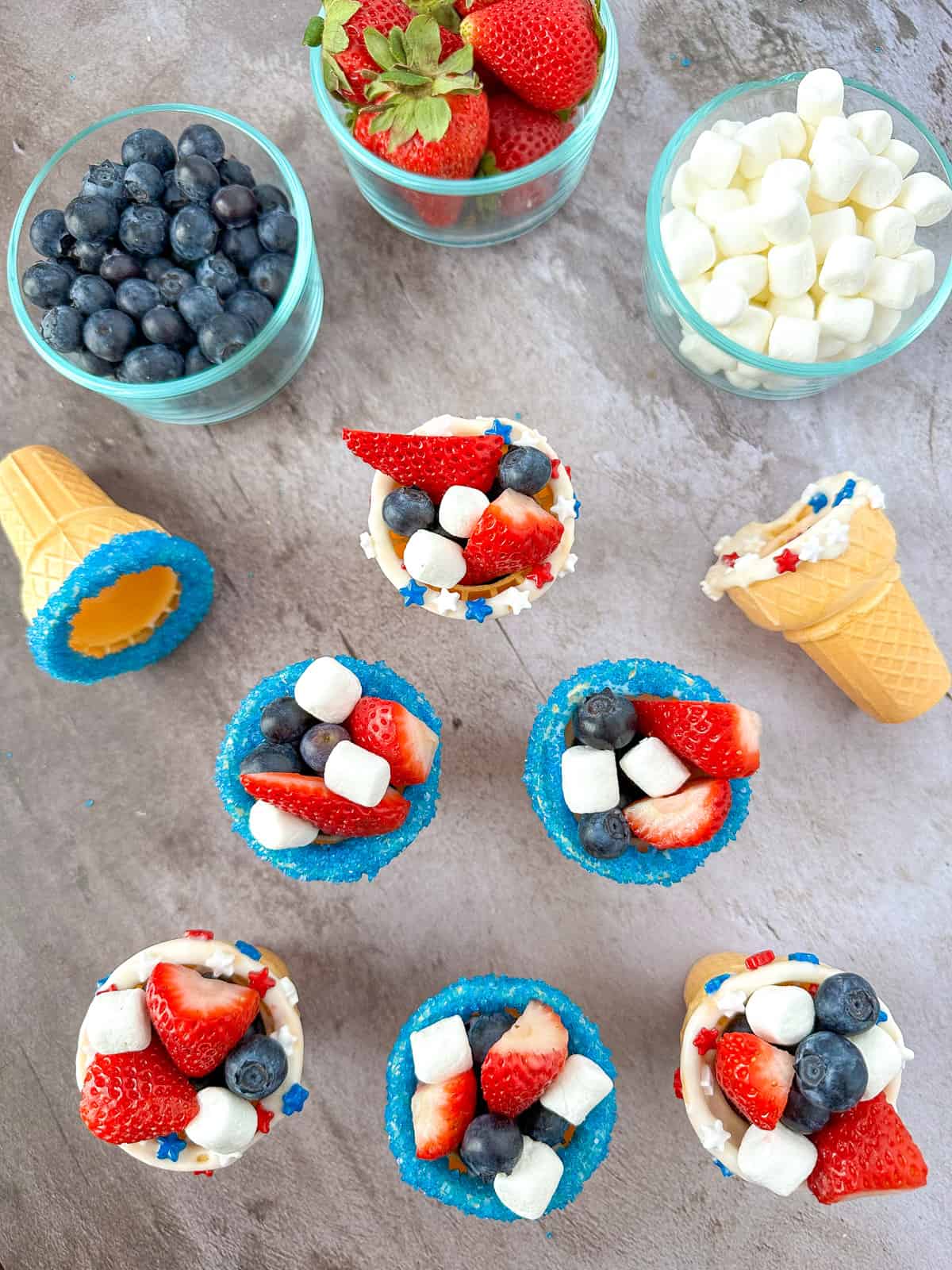 overhead view of a group of patriotic fruit ice cream cones with blueberries strawberries and marshmallows in clear cups in the background