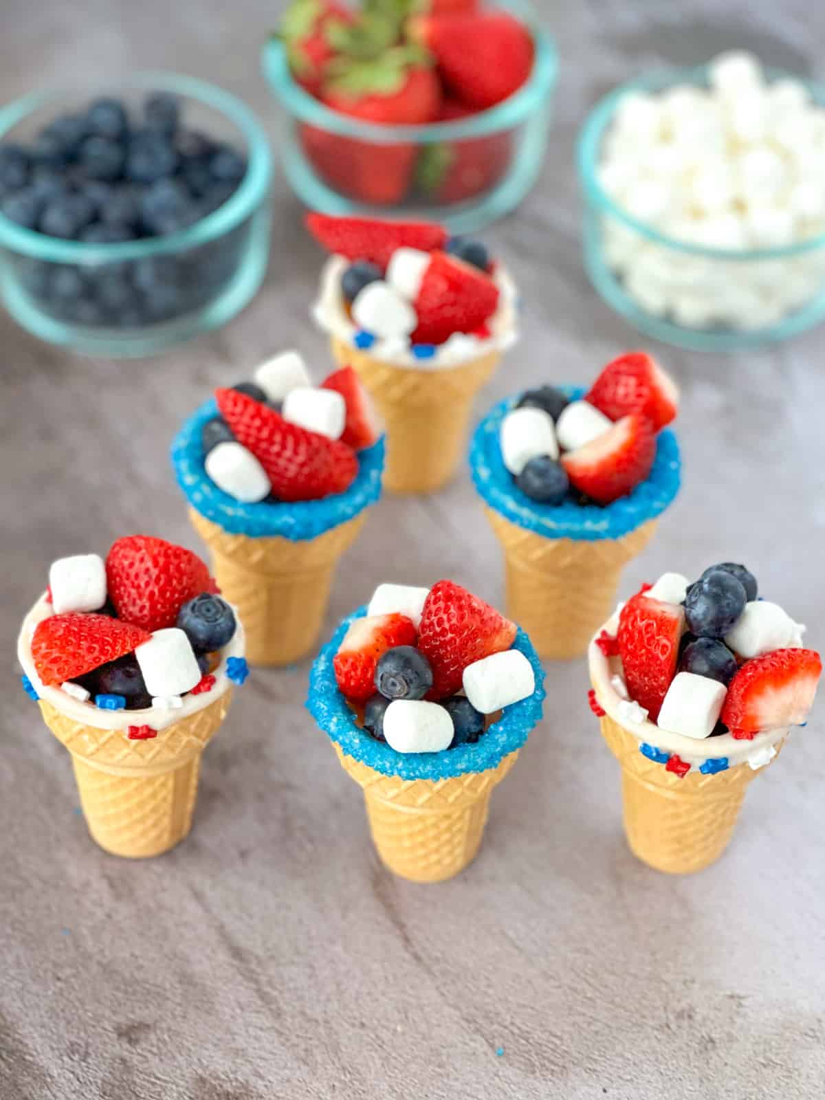 a group of patriotic fruit ice cream cones with blueberries strawberries and marshmallows in clear cups in the background