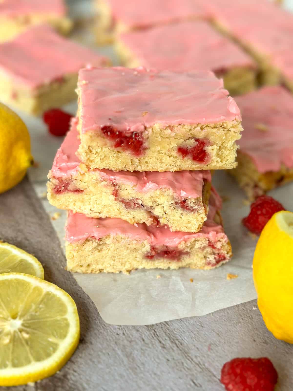 close up of a stack of 3 raspberry lemon blondies with more in the background plus lemons and raspberries