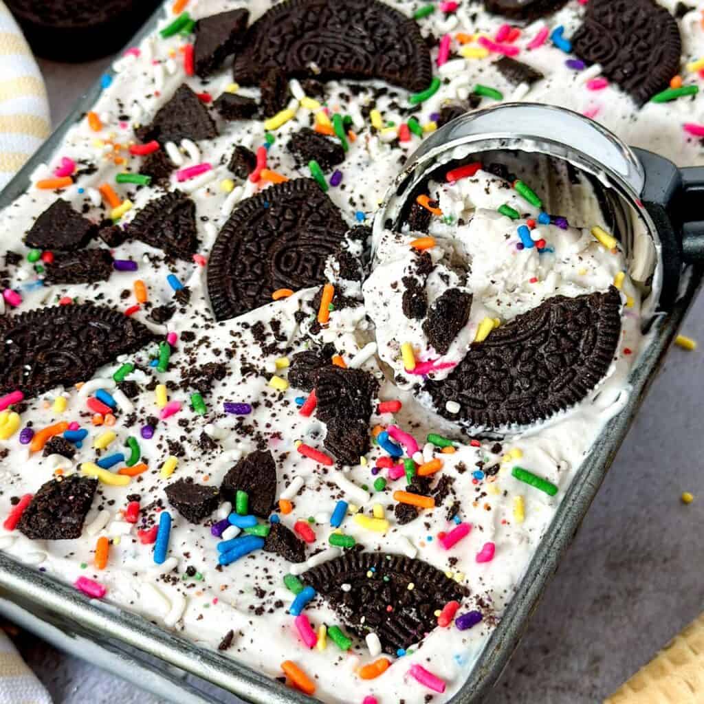 no churn birthday cake oreo ice cream in a pan being scooped out with a scoop with oreos and cones in the background