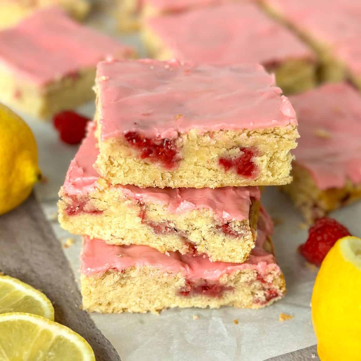 close up of a stack of 3 raspberry lemon blondies with more in the background plus lemons and raspberries