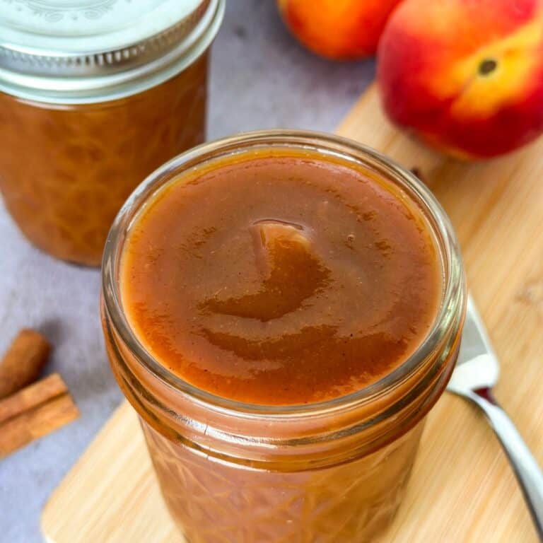 close up of homemade slow cooker peach butter in a mason jar with another full jar, cinnamon sticks, knife, and peaches in the background