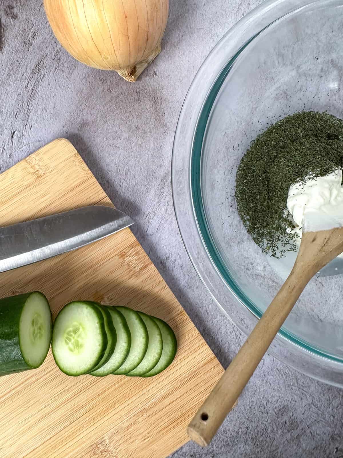 bowl of dressing, cutting board with a cucumber being sliced, and an onion on the side