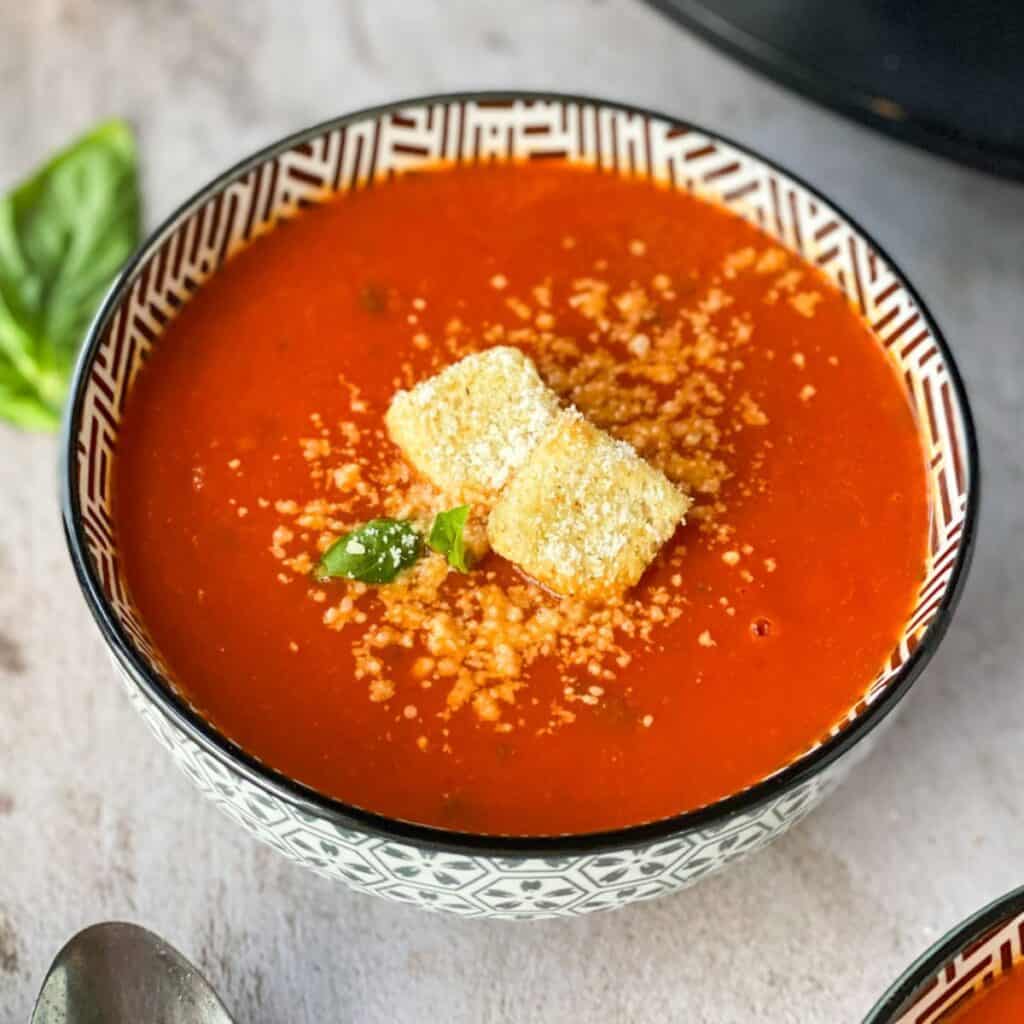 Easy Slow Cooker Tomato Basil Soup (with Canned Tomatoes) in a bowl with croutons and basil leaves with a crock pot in the background