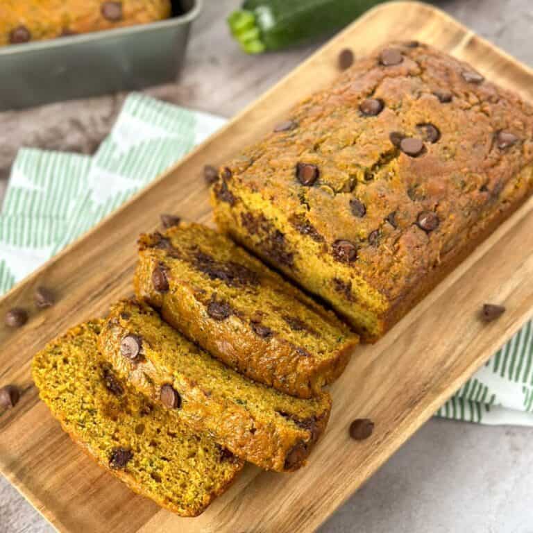 Pumpkin Zucchini Bread with Chocolate Chips being sliced on a cutting board with another loaf in a pan and a zucchini in the background