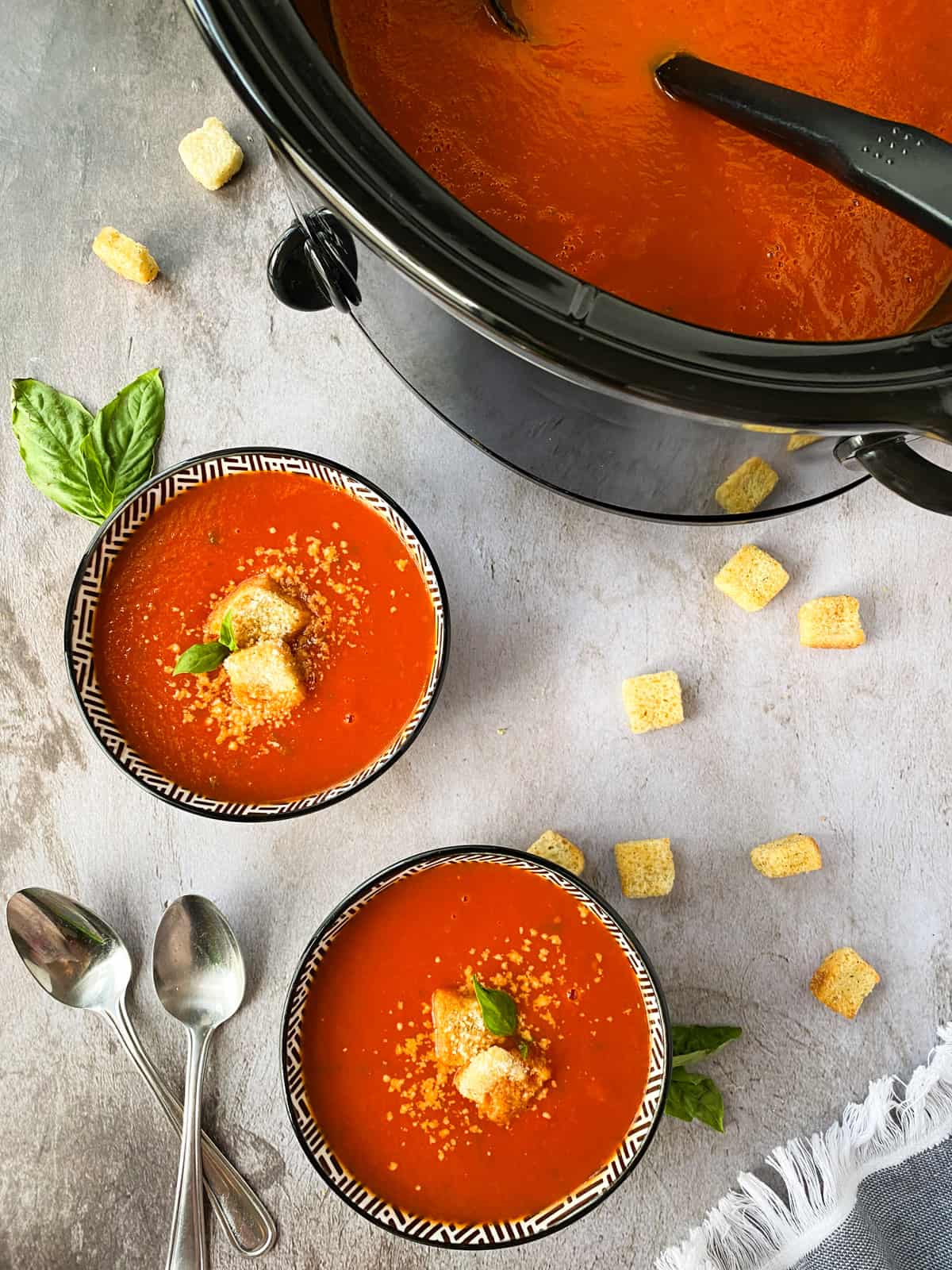 two bowls of Easy Slow Cooker Tomato Basil Soup with croutons, spoons, and basil leaves with a crock pot full of soup in the background