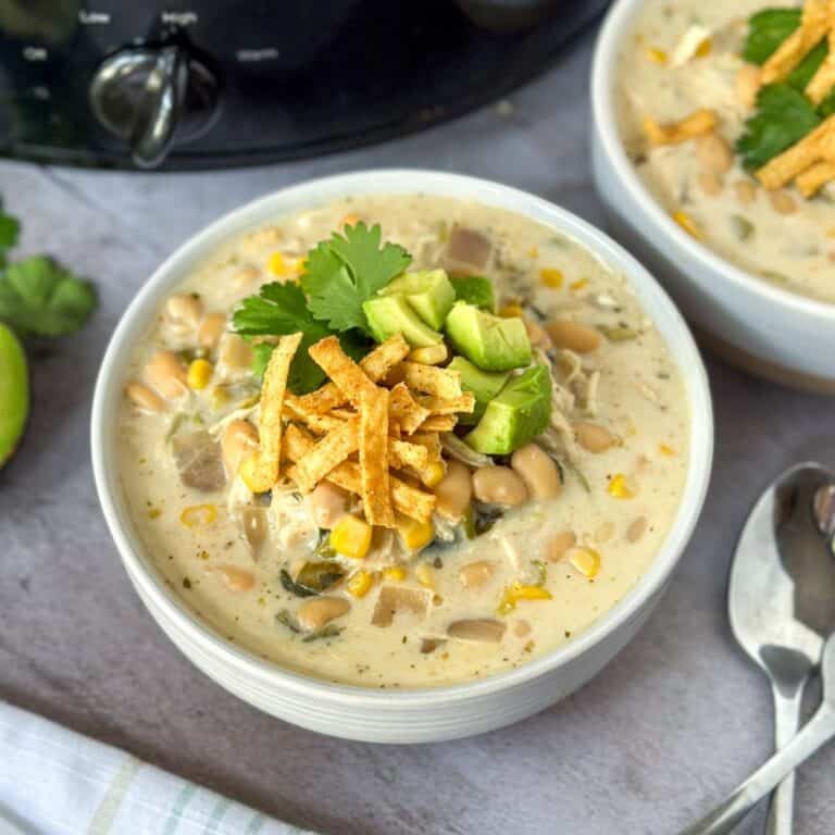 2 bowls of white chicken chili topped with avocado, cilantro, and tortilla strips with a crock pot next to it.