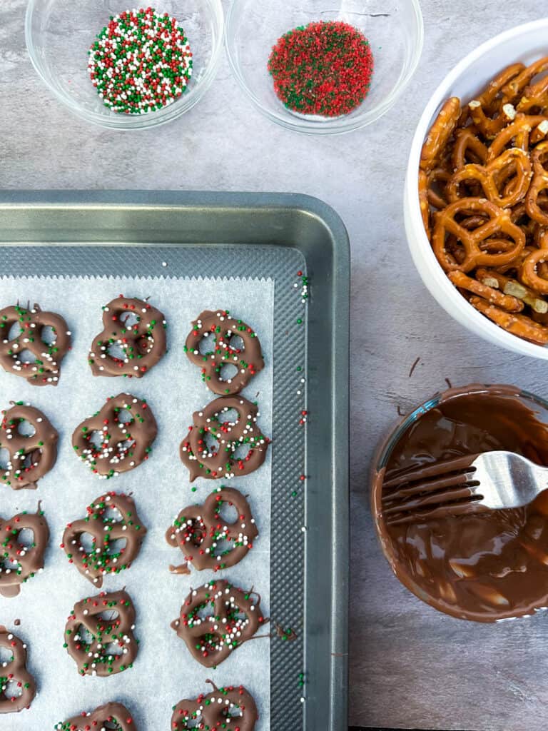 Chocolate covered pretzels on a baking sheet, a bowl of pretzels, two bowls of sprinkles, and a bowl of melted chocolate with a fork in it.