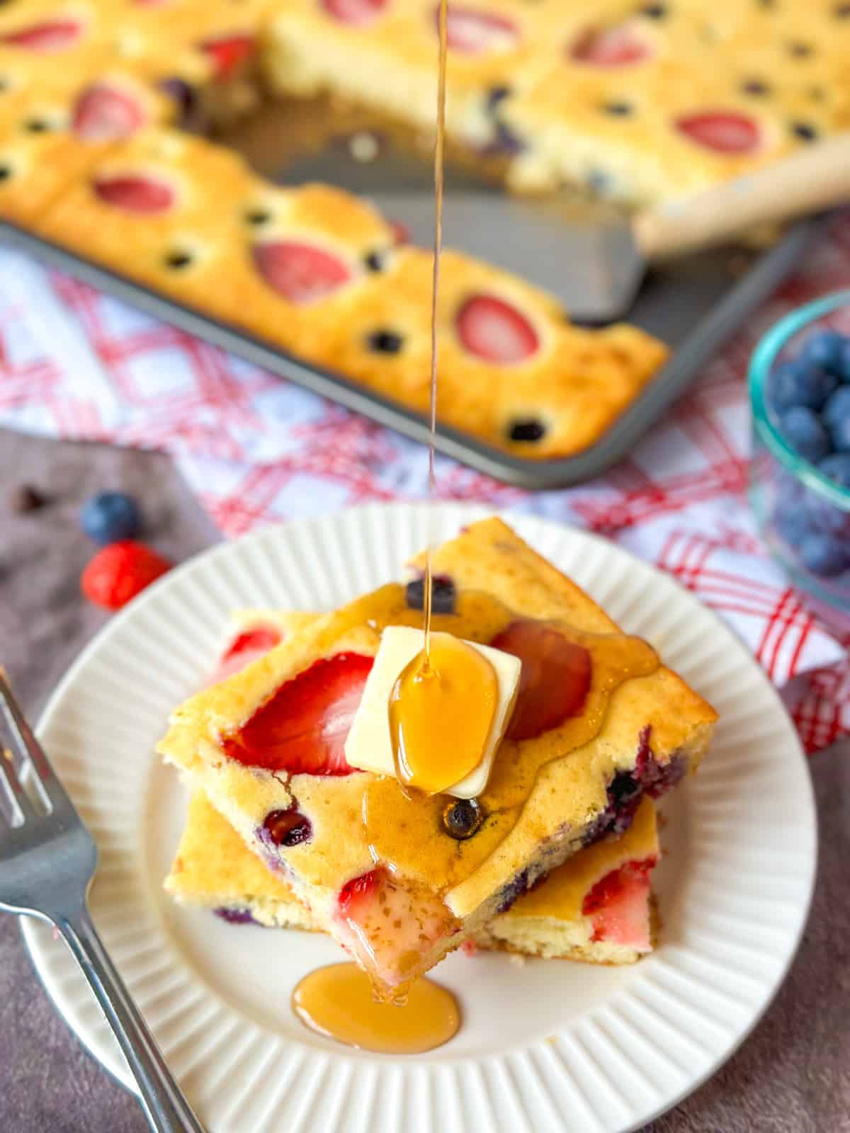 Sheet Pan Pancakes with two scooped on a plate with butter and maple syrup being poured over top with a bowl of blueberries on the side.