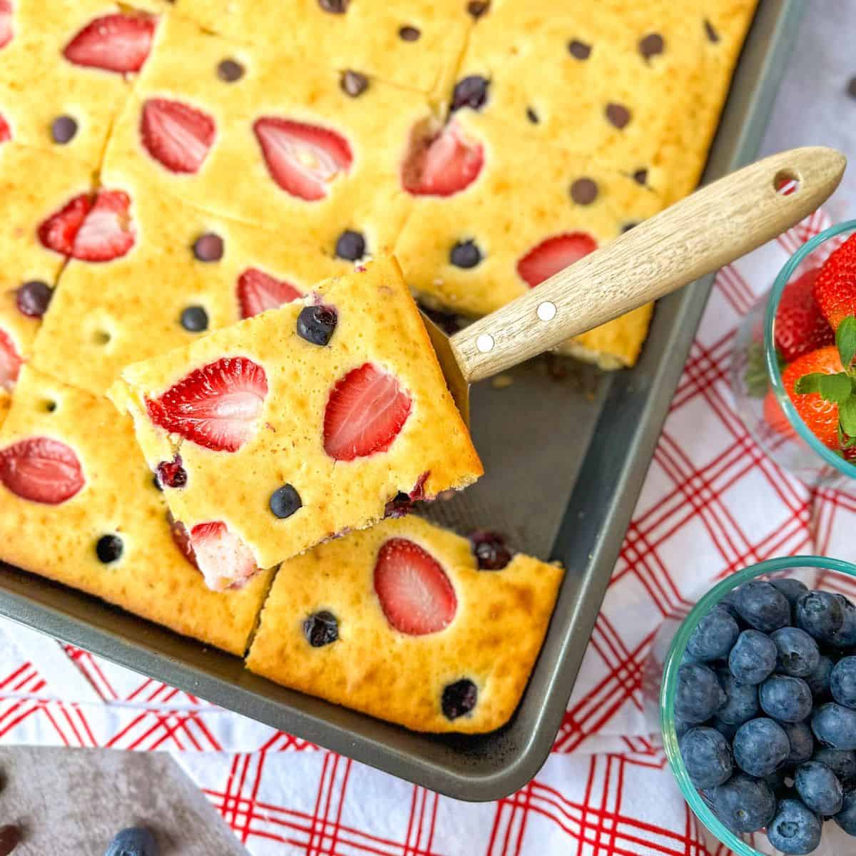 Sheet Pan Pancakes with one scooped on a spatula with a bowl of strawberries and blueberries on the side.