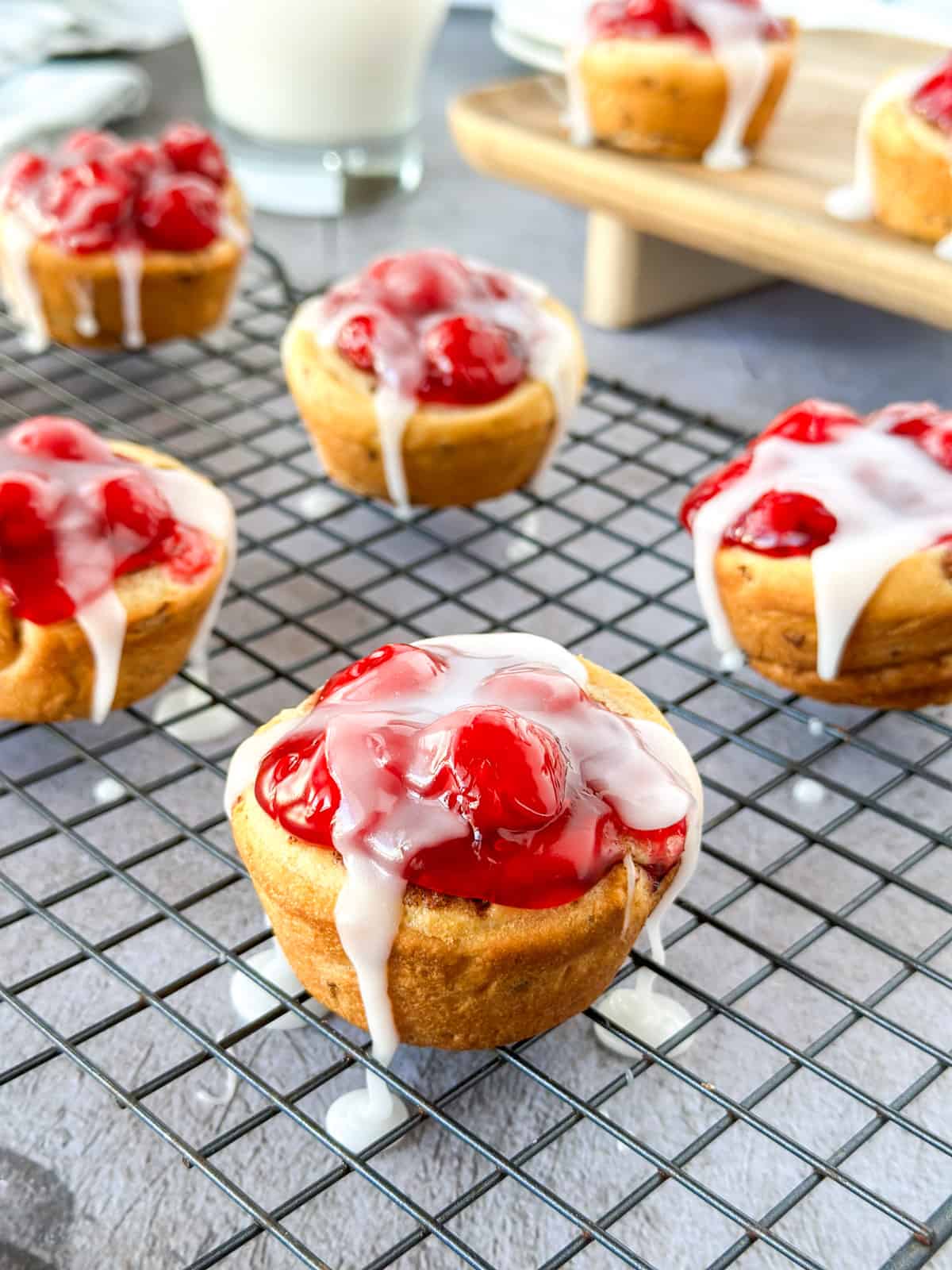 A cooling rack with cherry pie cups and icing drizzled over the top and a glass of milk behind it.