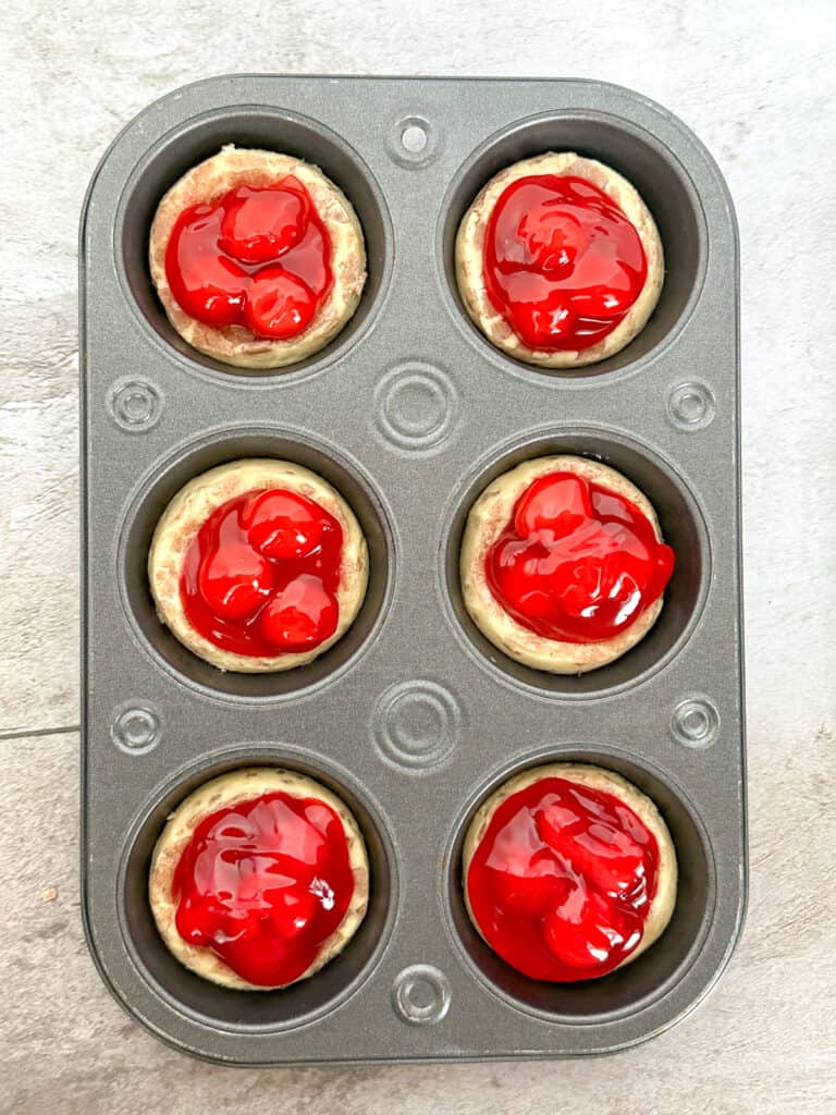 A muffin pan with cinnamon rolls and cherry pie filling in the center about to go in the oven.
