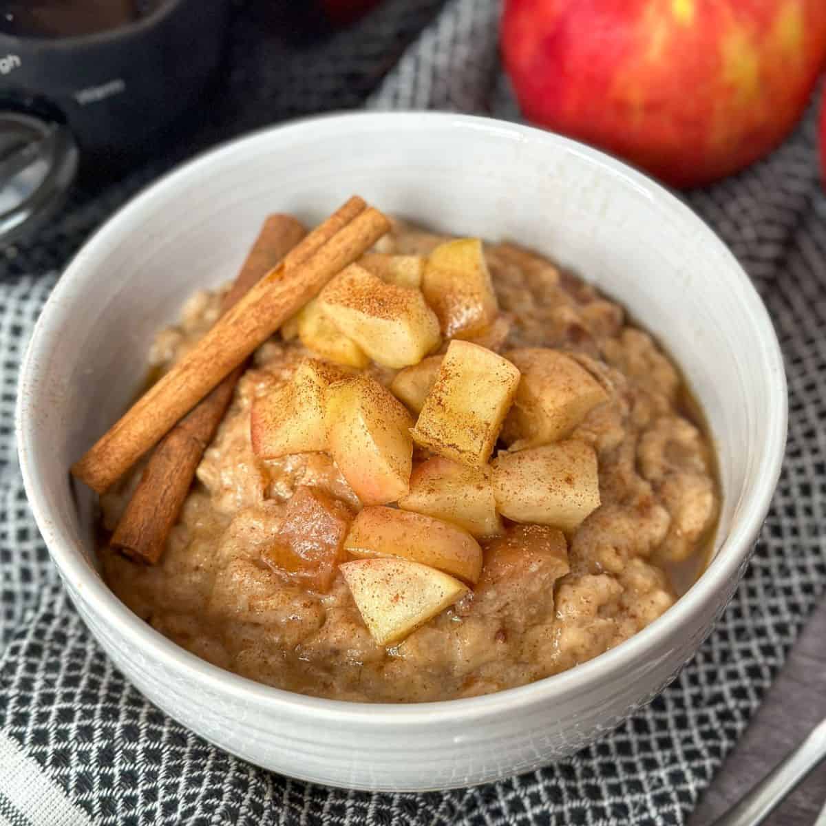 Bowl of Slow Cooker Apple Cinnamon Oatmeal with diced apples and cinnamon sticks on top with apples and a crock pot next to the bowl.