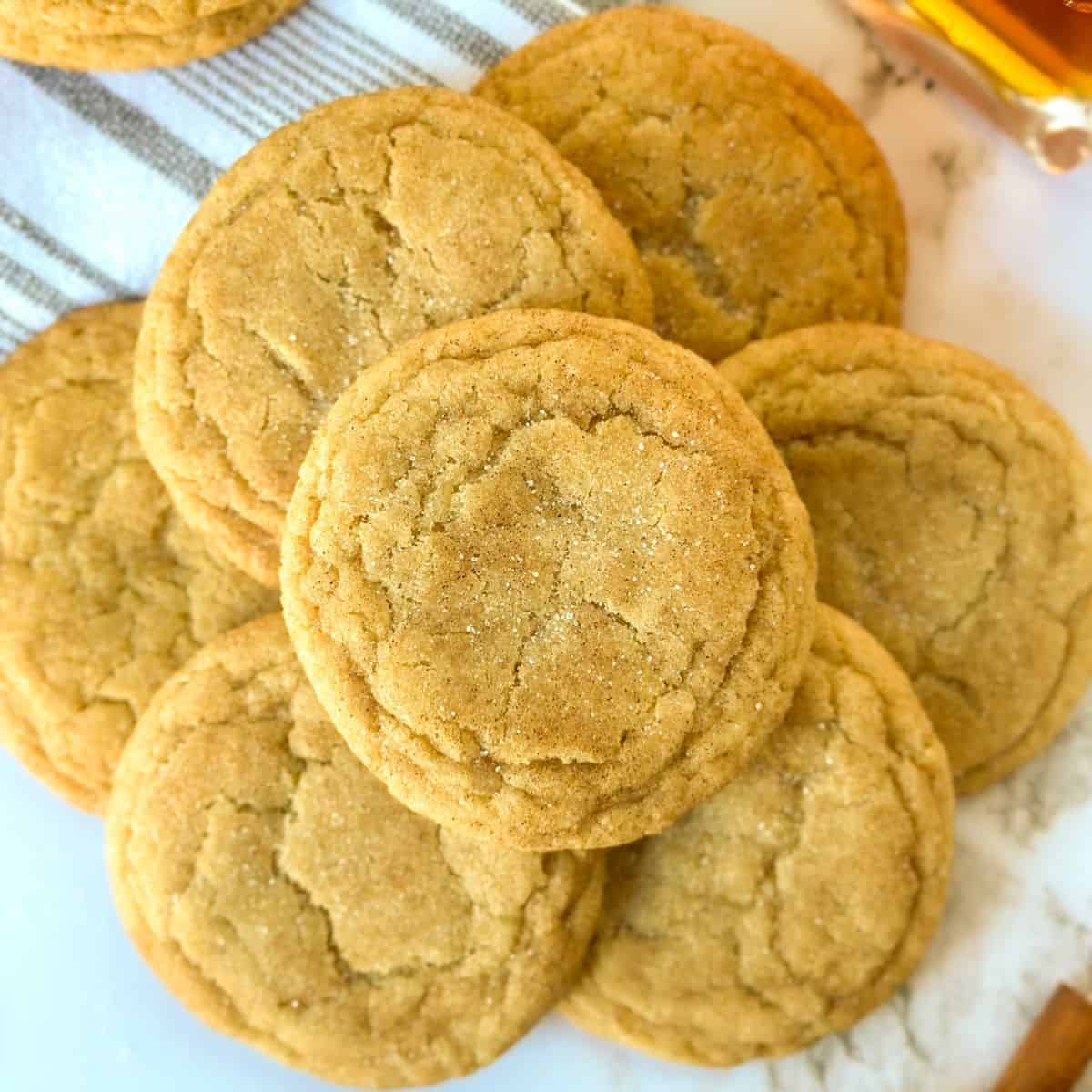 Maple Snickerdoodle cookies in a pile on the table with a bottle of maple syrup and cinnamon sticks next to them.