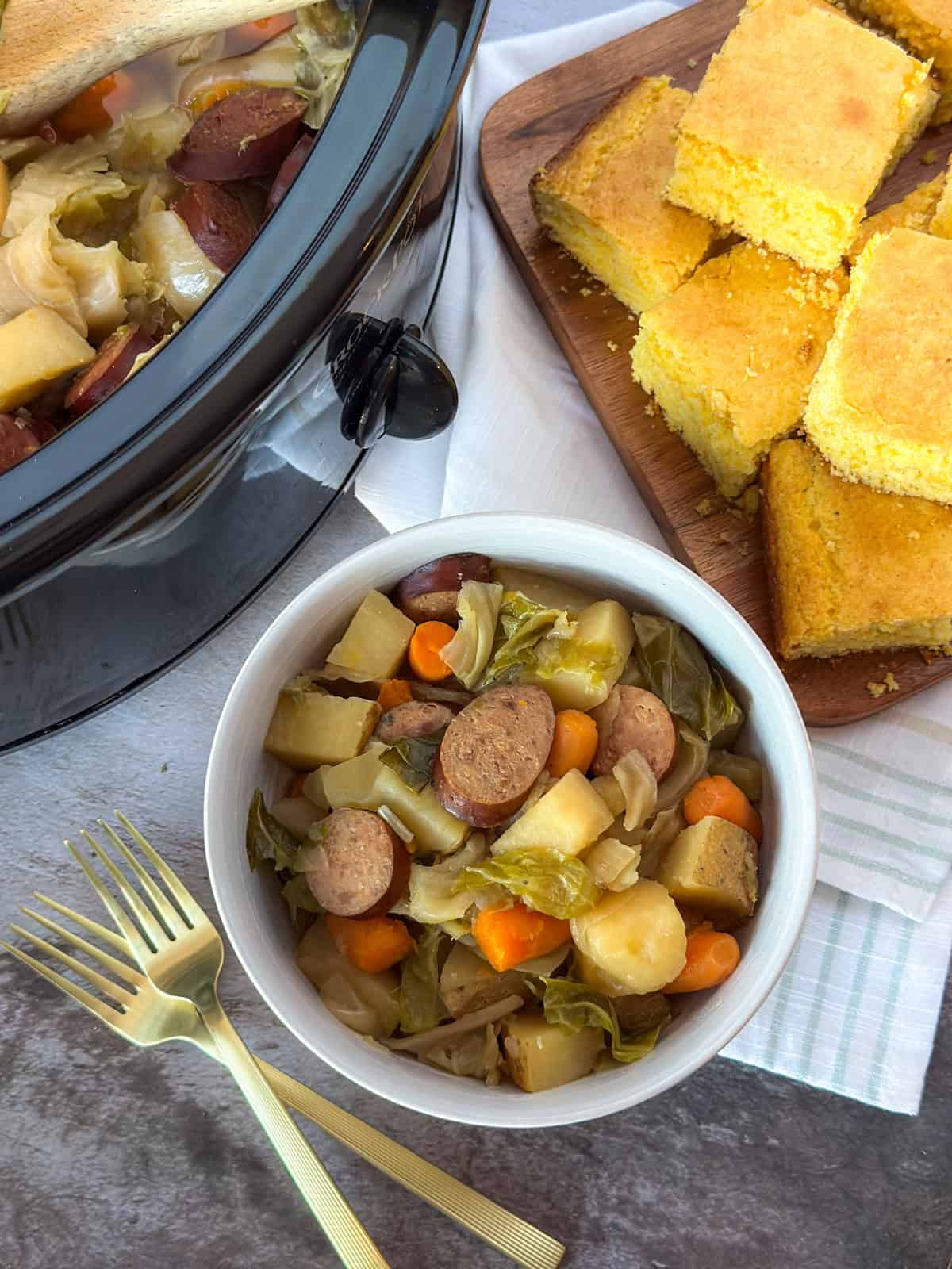 Crockpot Sausage, Potato, and Cabbage Recipe in a bowl on a table with a Crock Pot, 2 forks, and a cutting board with corn bread.