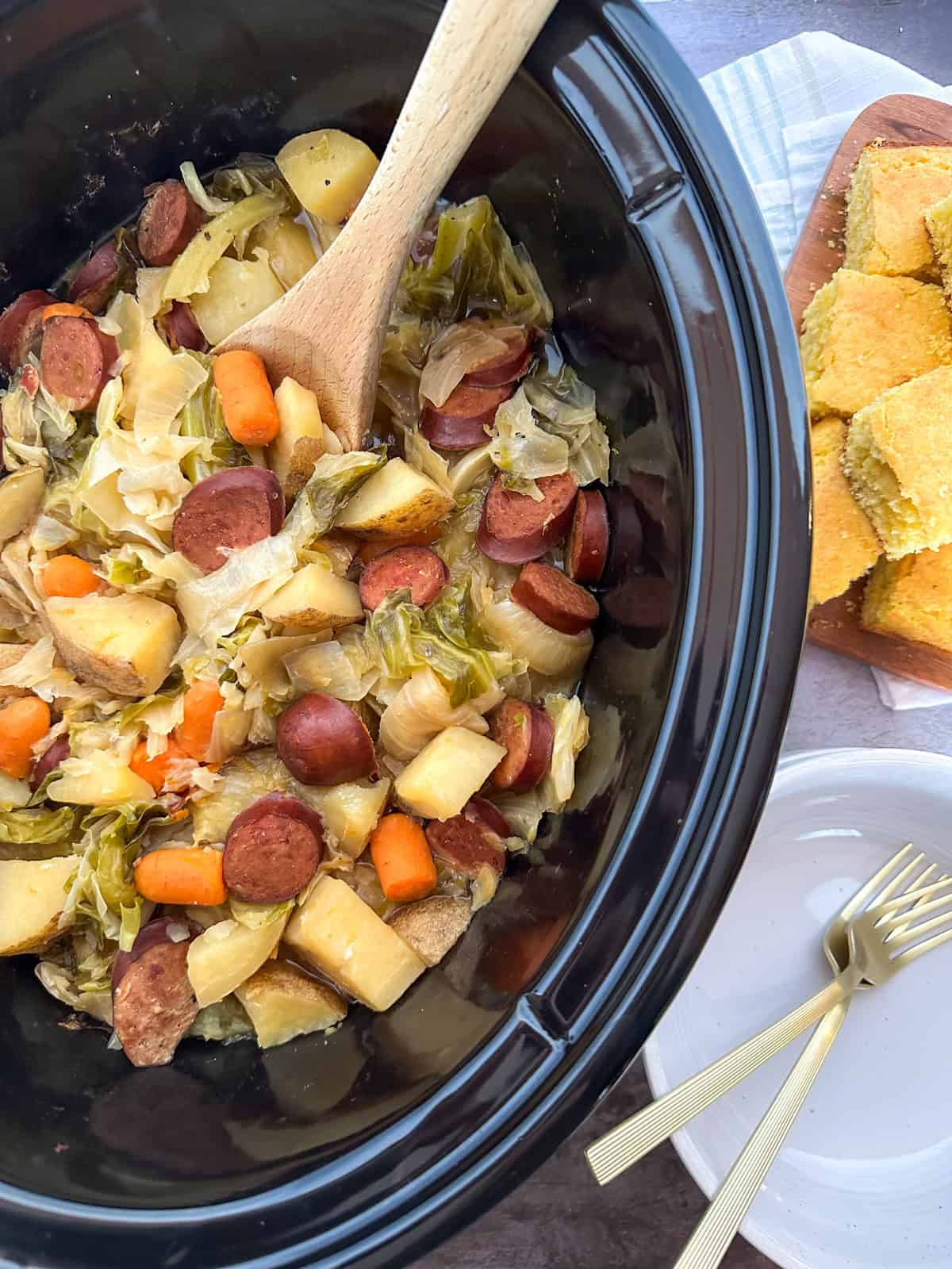 A crock pot filled with Sausage, Potato, and Cabbage on a table with corn bread and forks.