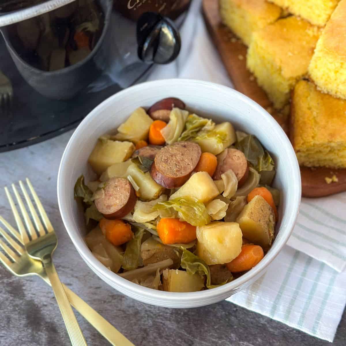 Crockpot Sausage, Potato, and Cabbage Recipe in a bowl on a table with a Crock Pot, 2 forks, and a cutting board with corn bread.