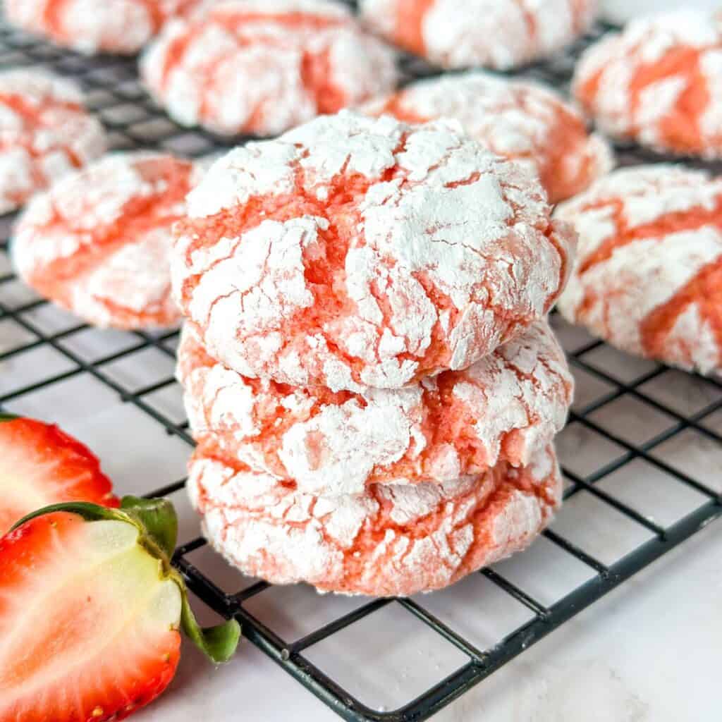 A stack of 3 Strawberry Crinkle Cool Whip Cookies on a cooling rack with cookies and cut strawberries surrounding them.