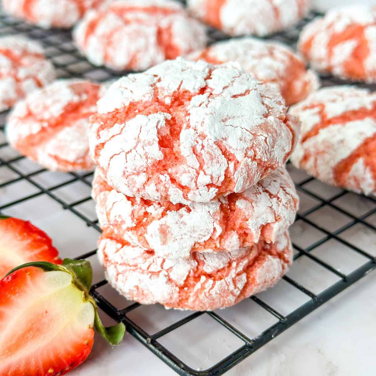 A stack of 3 Strawberry Crinkle Cool Whip Cookies on a cooling rack with cookies and cut strawberries surrounding them.