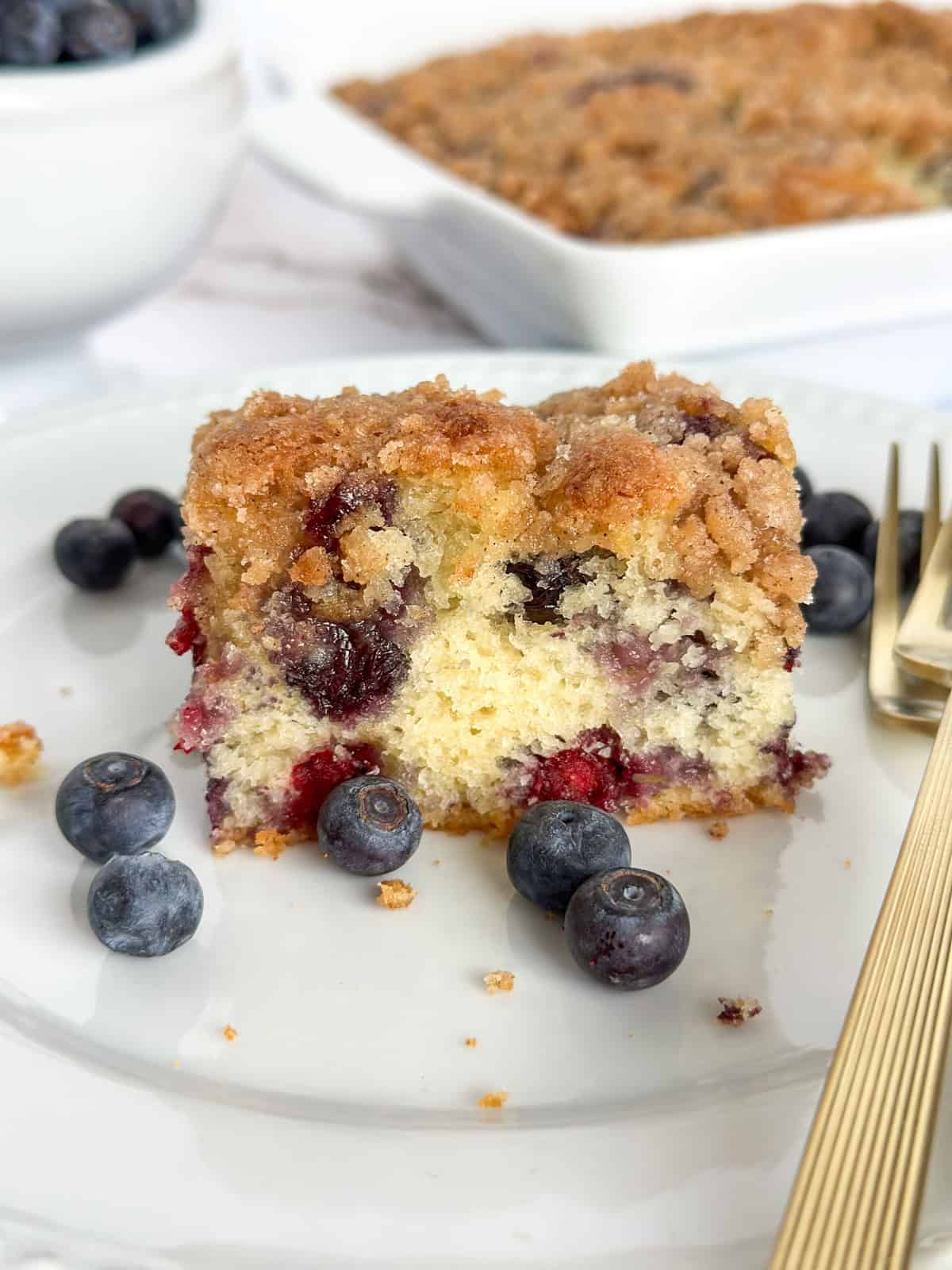 A slice of Old-Fashioned Blueberry Buckle Cake on a plate surrounded by blueberries and forks with a pan of cake behind the plate.