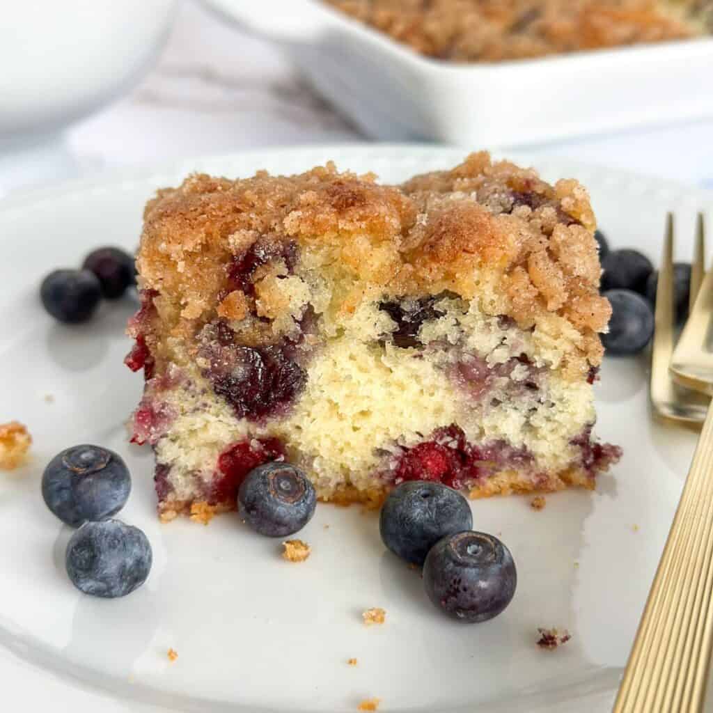A slice of Old-Fashioned Blueberry Buckle Cake on a plate surrounded by blueberries and forks with a pan of cake behind the plate.