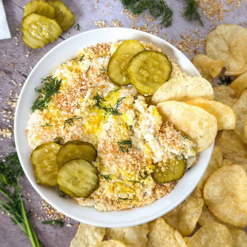 A bowl of Fried Dill Pickle Ranch Dip with chips in the bowl and next to it and pickles.