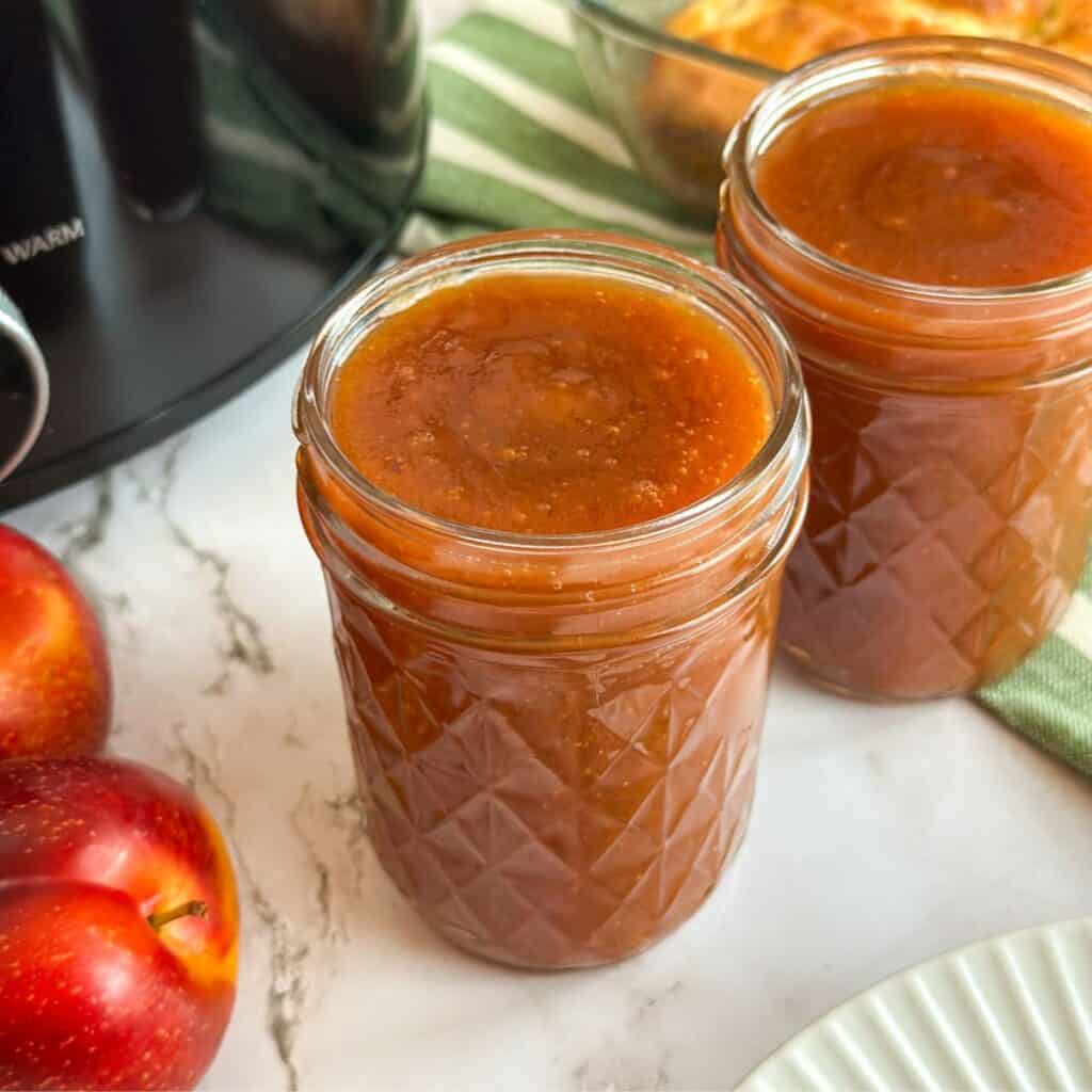 Two jars of plum butter with plums, a Crock Pot, a plate, and a pan of biscuits on the table.