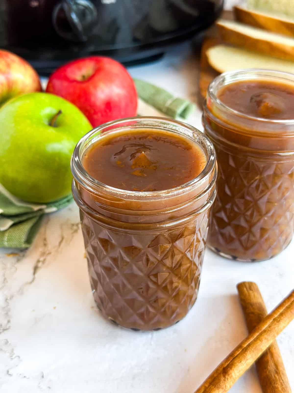 Two jars of Apple Butter with apples, cinnamon sticks, and a Crock Pot next to it.