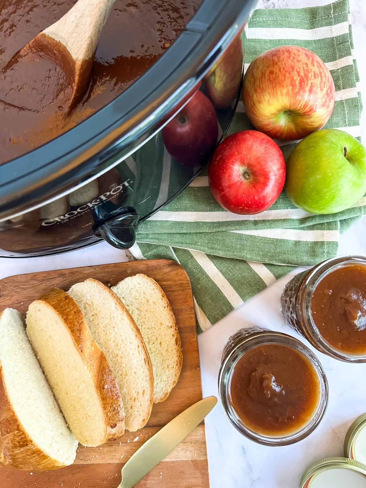 A Crock Pot filled with apple butter, two jars of apple butter, slices of bread on a cutting board, and apples on a table.