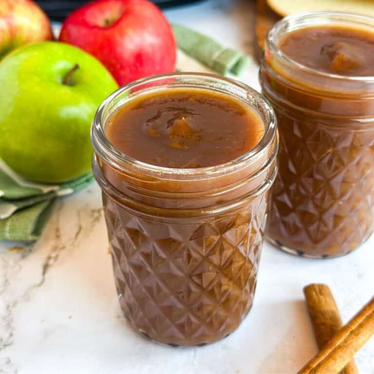 Two jars of Apple Butter with apples, cinnamon sticks, and a Crock Pot next to it.