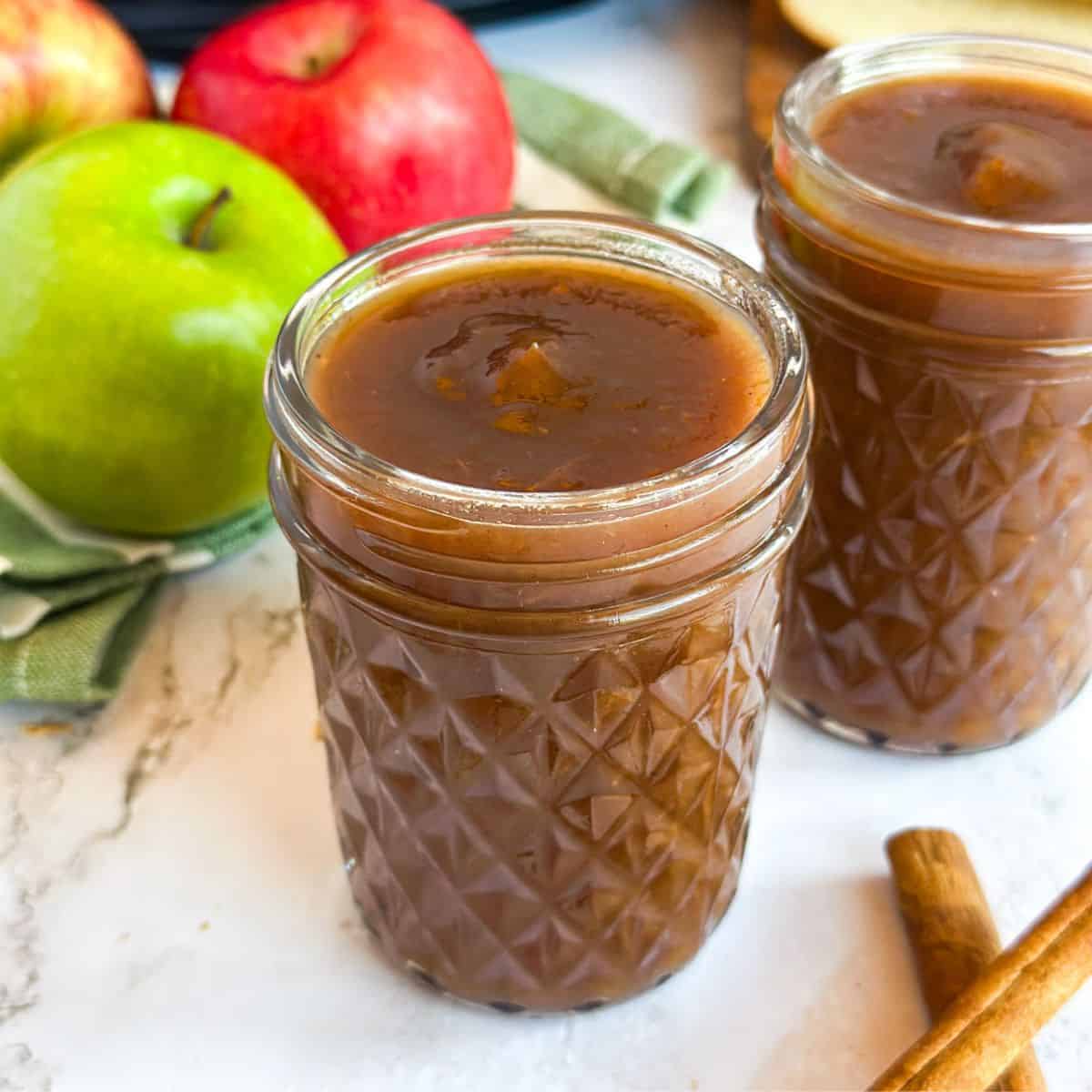 Two jars of Apple Butter with apples, cinnamon sticks, and a Crock Pot next to it.