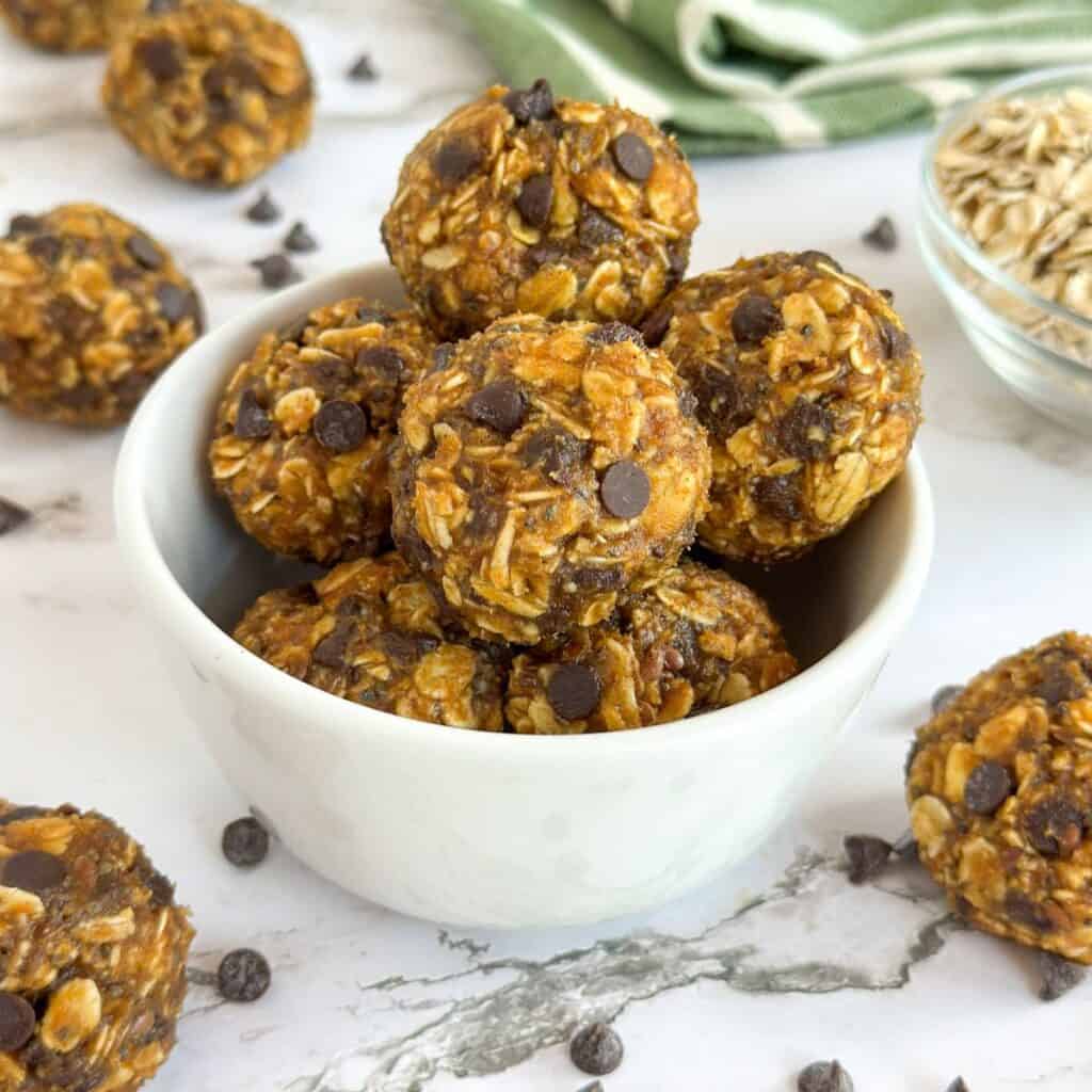 No-Bake Pumpkin Spice Energy Balls in a cup on a table with more balls, a cloth napkin, and a bowl of oats surrounding it.