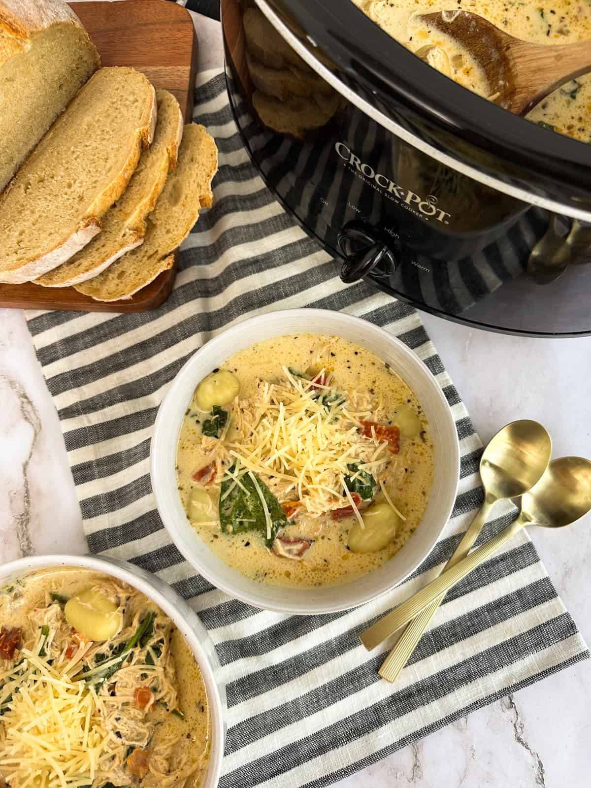 A bowl of Slow Cooker Marry Me Chicken Gnocchi Soup with another bowl, a slow cooker, spoons, and a loaf of bread next to it.