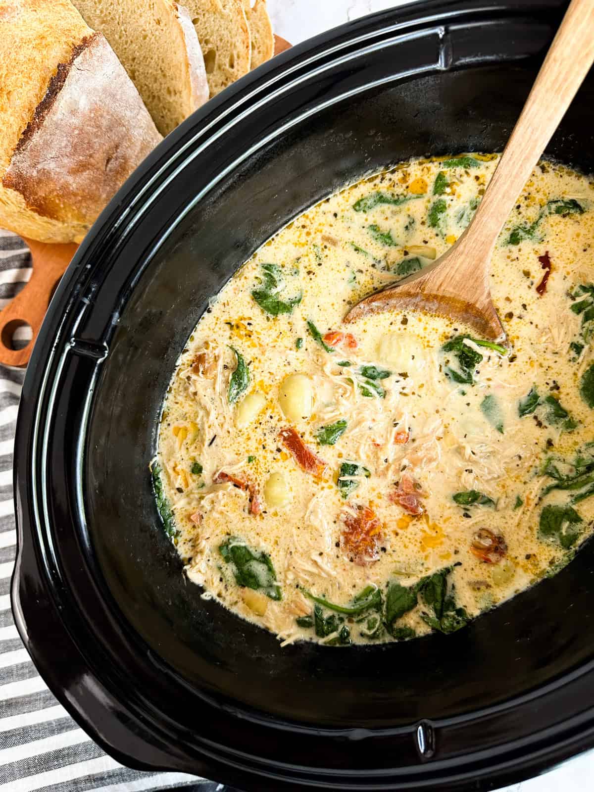 A Crock Pot filled with Marry Me Chicken Gnocchi Soup with a cutting board with bread next to it.